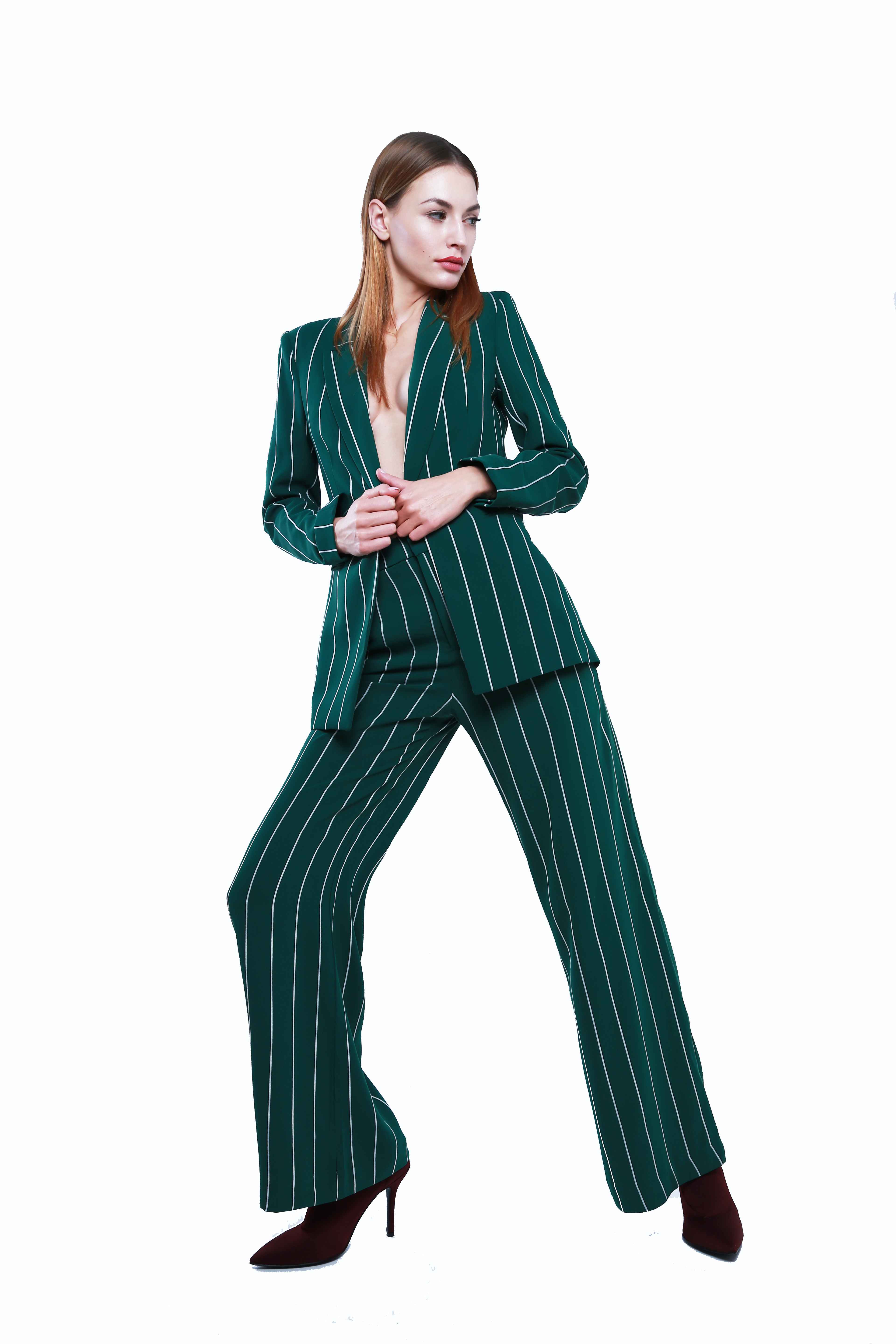 Kobieta w zielonym, prążkowanym garniturze pozuje na białym tle, z delikatnym uśmiechem i bordowymi botkami na obcasie.
