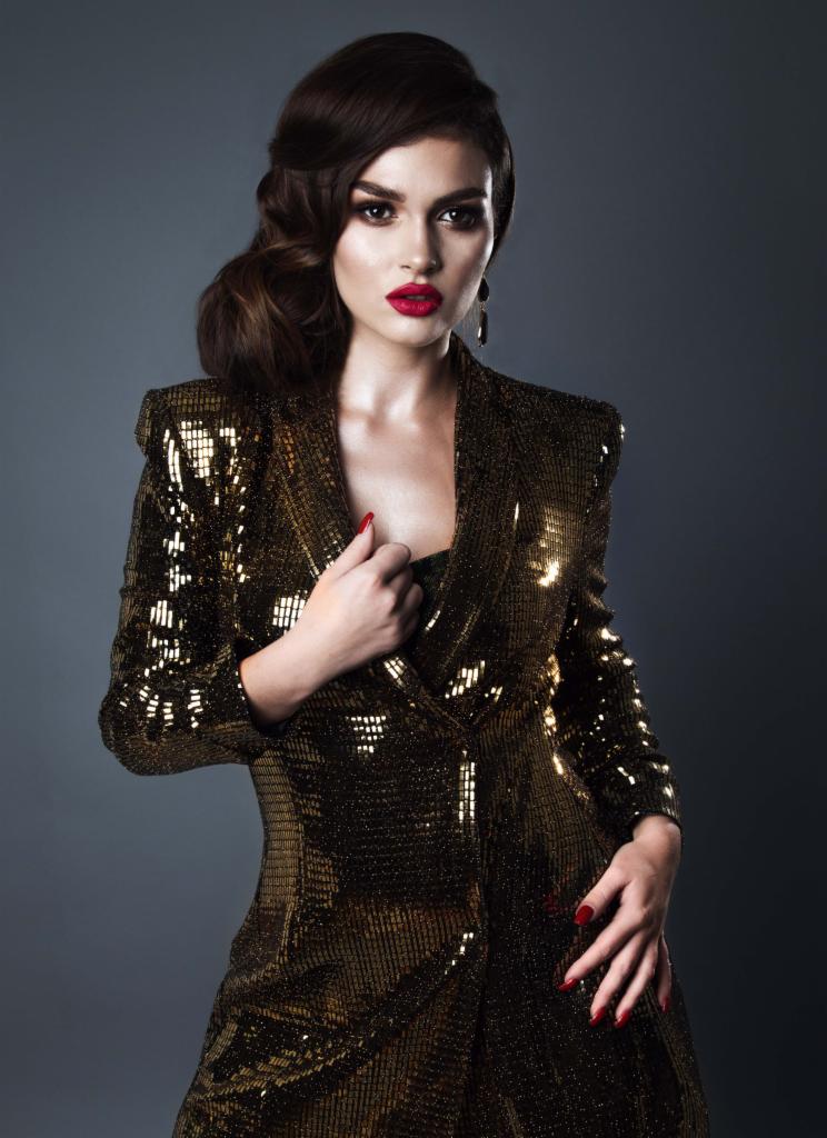 Kobieta w cekinowym złotym żakiecie, z czerwonymi ustami i upiętymi włosami, pozuje na ciemnoszarym tle.