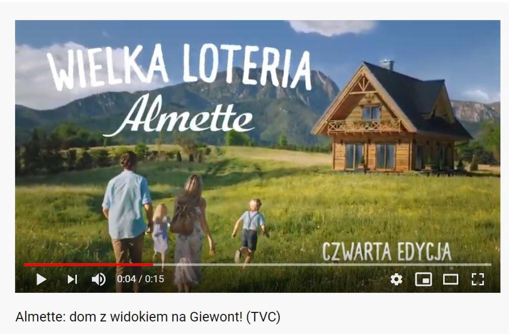 Rodzina spaceruje po łące w kierunku drewnianego domu z widokiem na góry, reklama loterii Almette.