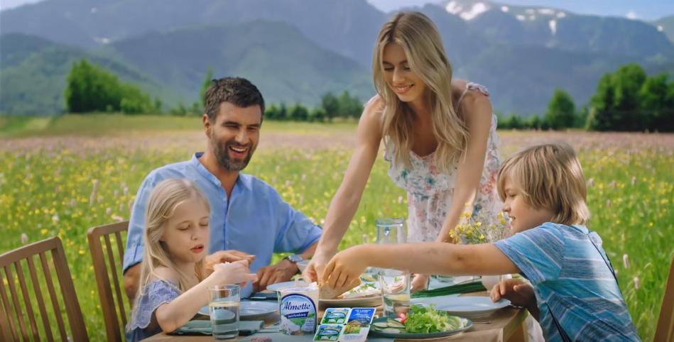 Rodzina je śniadanie na stole w plenerze, w tle łąka i góry, na stole widoczny serek Almette.