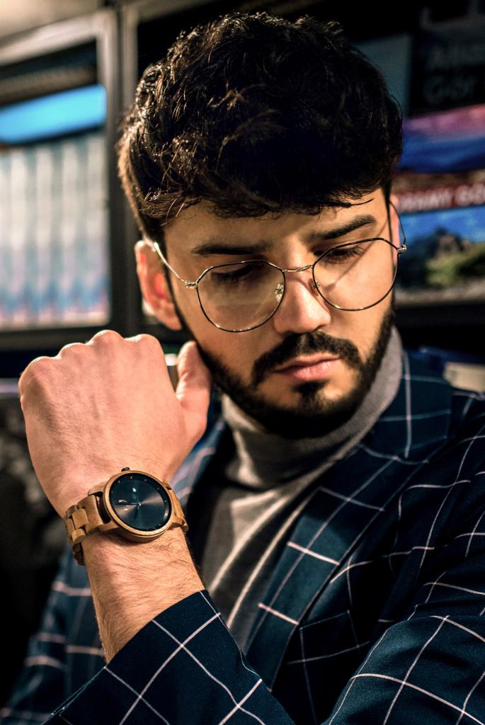 Mężczyzna z brodą w okularach i marynarce w kratę, prezentujący drewniany zegarek na ręce.