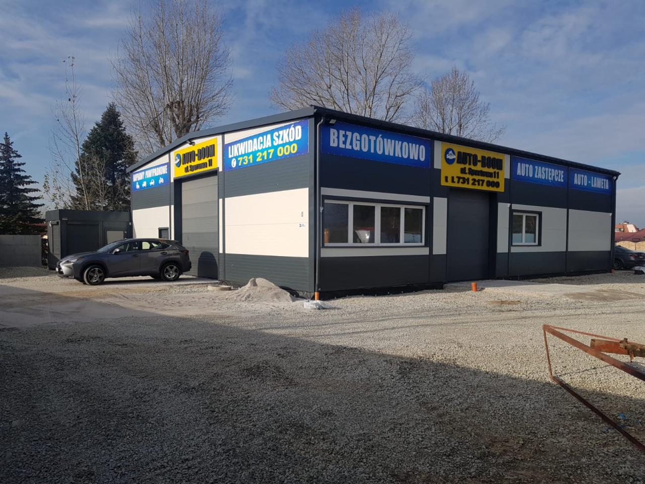 Nowo wybudowany warsztat samochodowy Auto-Boom z reklamami usług likwidacji szkód i aut zastępczych, szary samochód zaparkowany przed budynkiem.