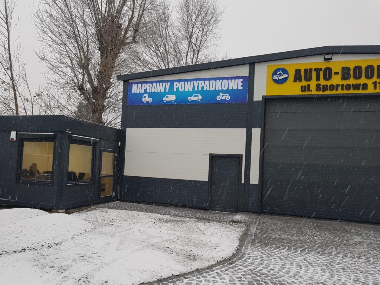 Zimowy widok warsztatu samochodowego z szyldem 'Naprawy powypadkowe' i żółtym logo 'AUTO-BOOK', kobieta widoczna w oknie biura.
