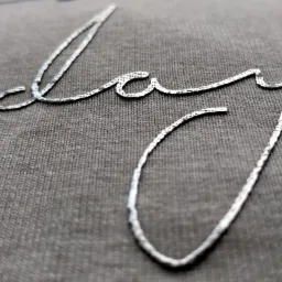 Szary t-shirt z wyhaftowanym srebrną nicią napisem w kursywie, widoczne zbliżenie na fakturę materiału i detale ściegu.