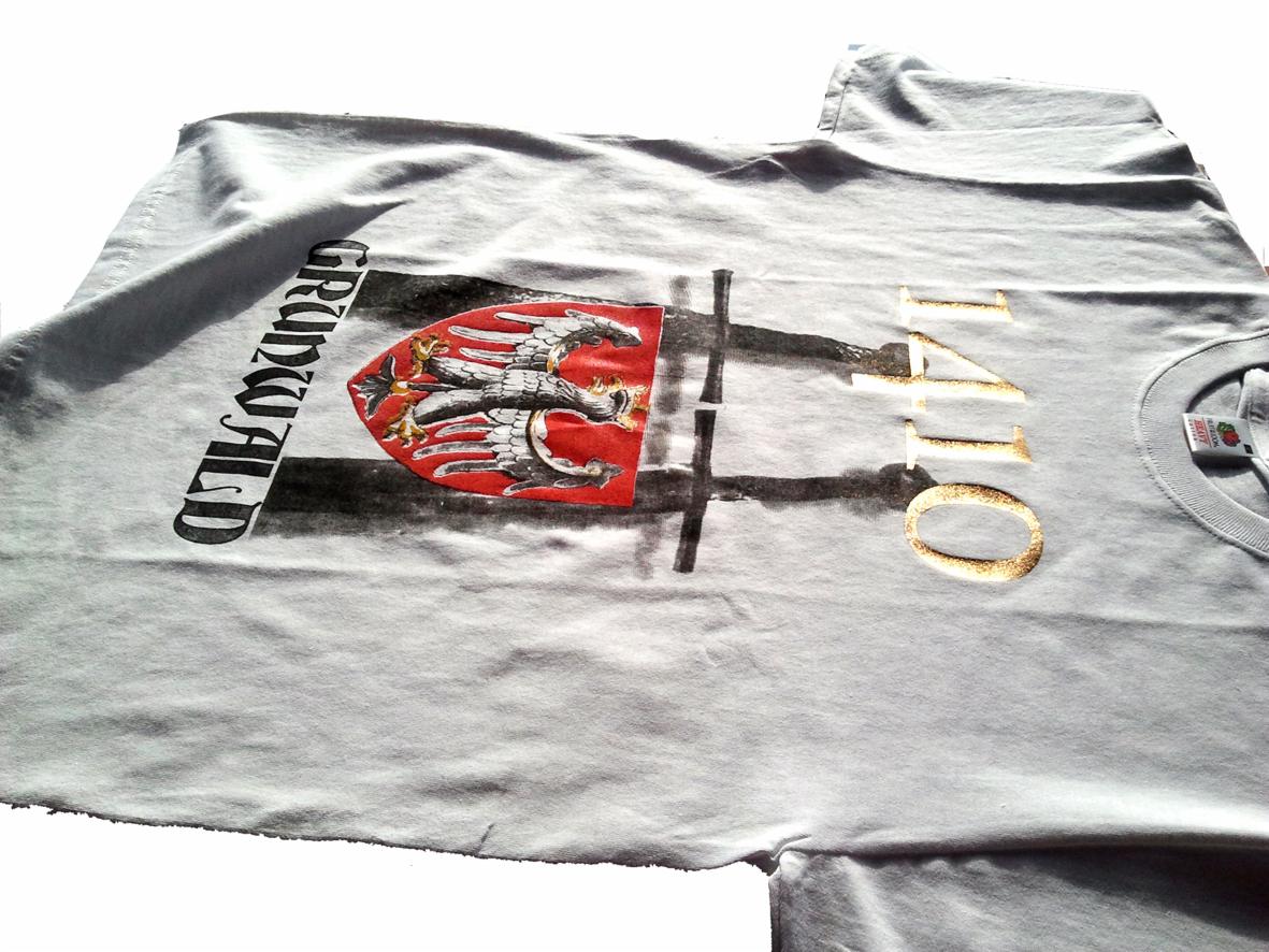 Jasnoszara koszulka z nadrukiem herbu z orłem na czerwonej tarczy, napisem 'Grunwald' i datą '1410' w złotym kolorze, leżąca na białym tle.