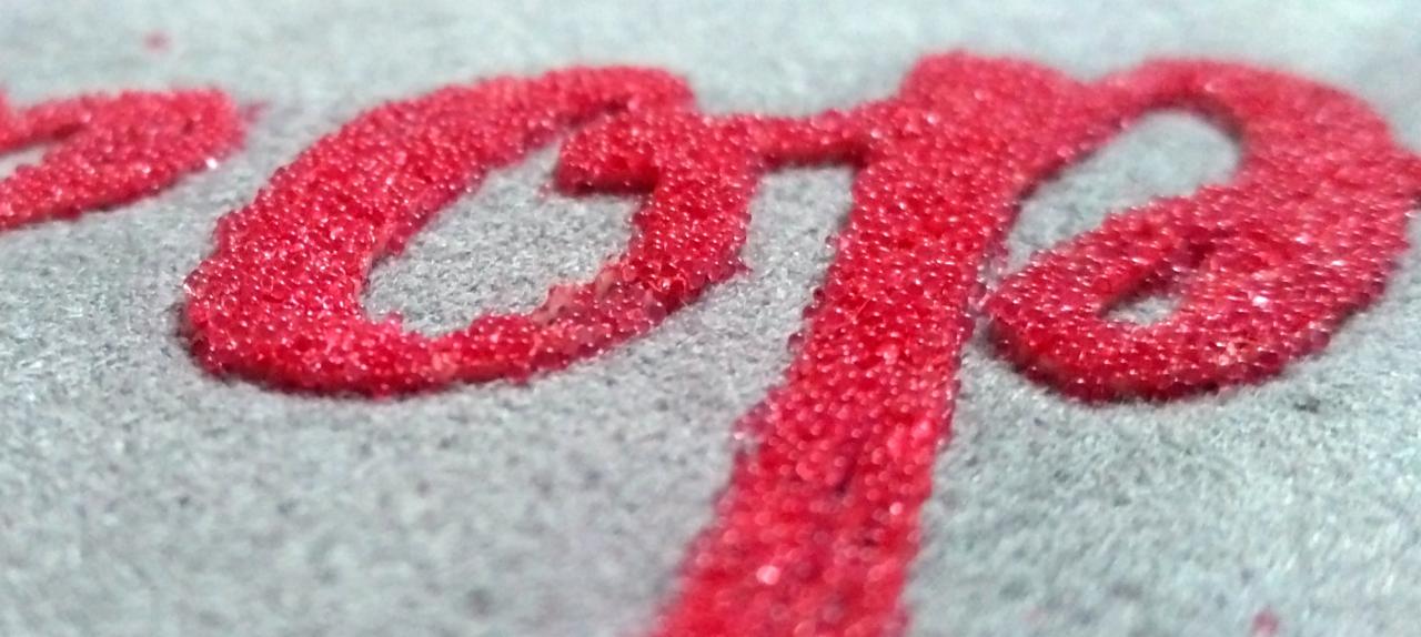 Czerwony, teksturowany napis na szarym materiale, widoczne z bliska, z efektem trójwymiarowości.