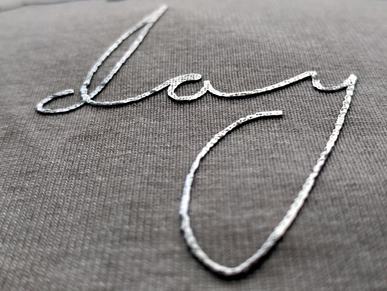 Szary t-shirt z wyhaftowanym srebrną nicią napisem w kursywie, widoczne zbliżenie na fakturę materiału i detale ściegu.