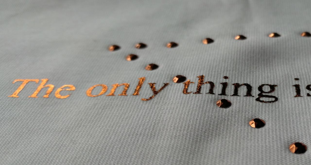 Jasnoniebieska tkanina z napisem 'The only thing is' wykonanym złotą czcionką i ozdobiona brązowymi dżetami