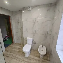 Nowoczesna łazienka z białymi płytkami imitującymi marmur, drewnopodobną podłogą, toaletą i bidetem. Widoczny fragment kabiny prysznicowej i otwarte drzwi do innego pomieszczenia.