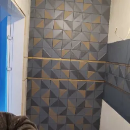 Ściana łazienki wyłożona dekoracyjnymi, trójwymiarowymi płytkami w odcieniach szarości i brązu, z widocznymi fragmentami instalacji hydraulicznej oraz fragmentem ościeżnicy drzwiowej zabezpieczonej...