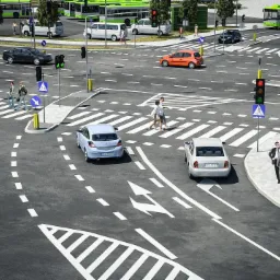 Szeroki plan skrzyżowania z przejściami dla pieszych, sygnalizacją świetlną i ruchem ulicznym, w słoneczny dzień, z widocznymi znakami drogowymi i liniami poziomymi.