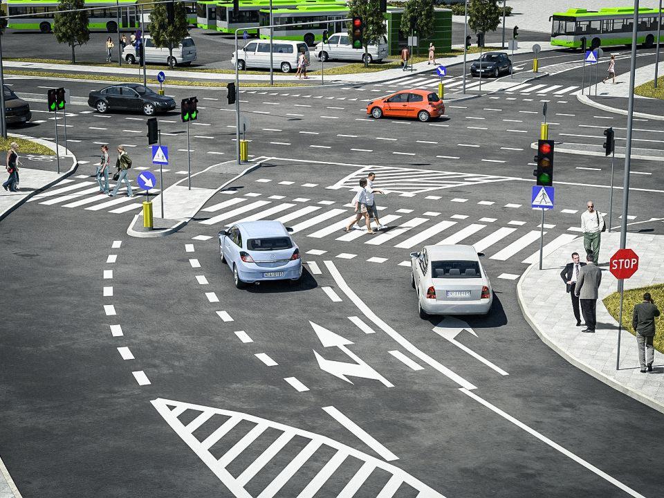 Szeroki plan skrzyżowania z przejściami dla pieszych, sygnalizacją świetlną i ruchem ulicznym, w słoneczny dzień, z widocznymi znakami drogowymi i liniami poziomymi.