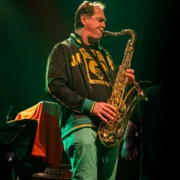 Saksofonista w czarnej kurtce z żółto-zielonymi detalami gra na saksofonie podczas występu na scenie, oświetlony zielonym światłem.