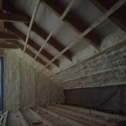 Poddasze z drewnianą konstrukcją dachu, częściowo pokryte jasną, spienioną izolacją, widoczna drabina i folia ochronna.