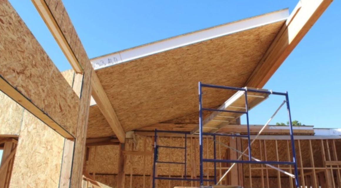 Szkielet drewniany domu w trakcie budowy, widoczne płyty OSB i rusztowanie na tle błękitnego nieba.
