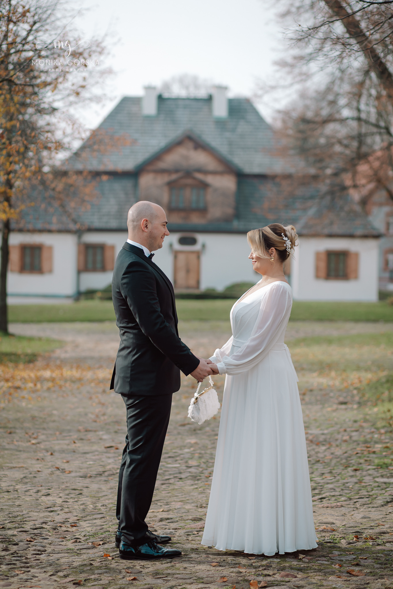 Para młoda trzymająca się za ręce na tle dworku z jesiennymi drzewami. Panna młoda w białej sukni z długim rękawem, pan młody w czarnym smokingu.