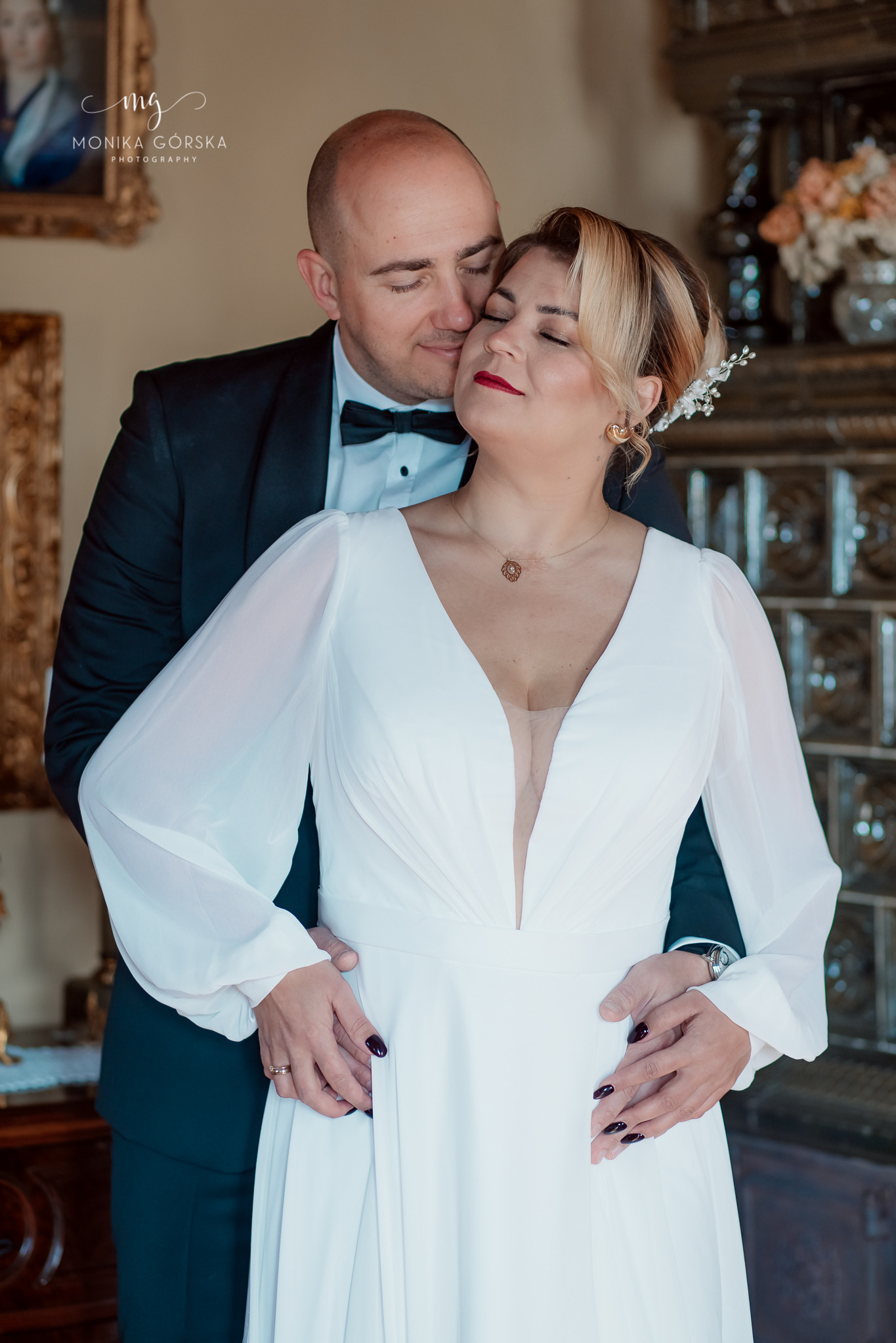 Uśmiechnięta para młoda w objęciach na tle zdobionego pieca kaflowego. Panna młoda w białej sukni z długimi rękawami, pan młody w czarnym smokingu.