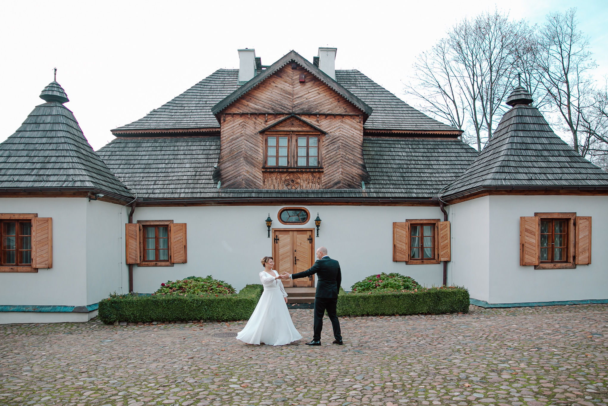 Para młoda w tańcu na brukowanym dziedzińcu przed tradycyjnym, drewnianym dworkiem z charakterystycznym dachem i okiennicami. Ujęcie z daleka, pełna sylwetka.