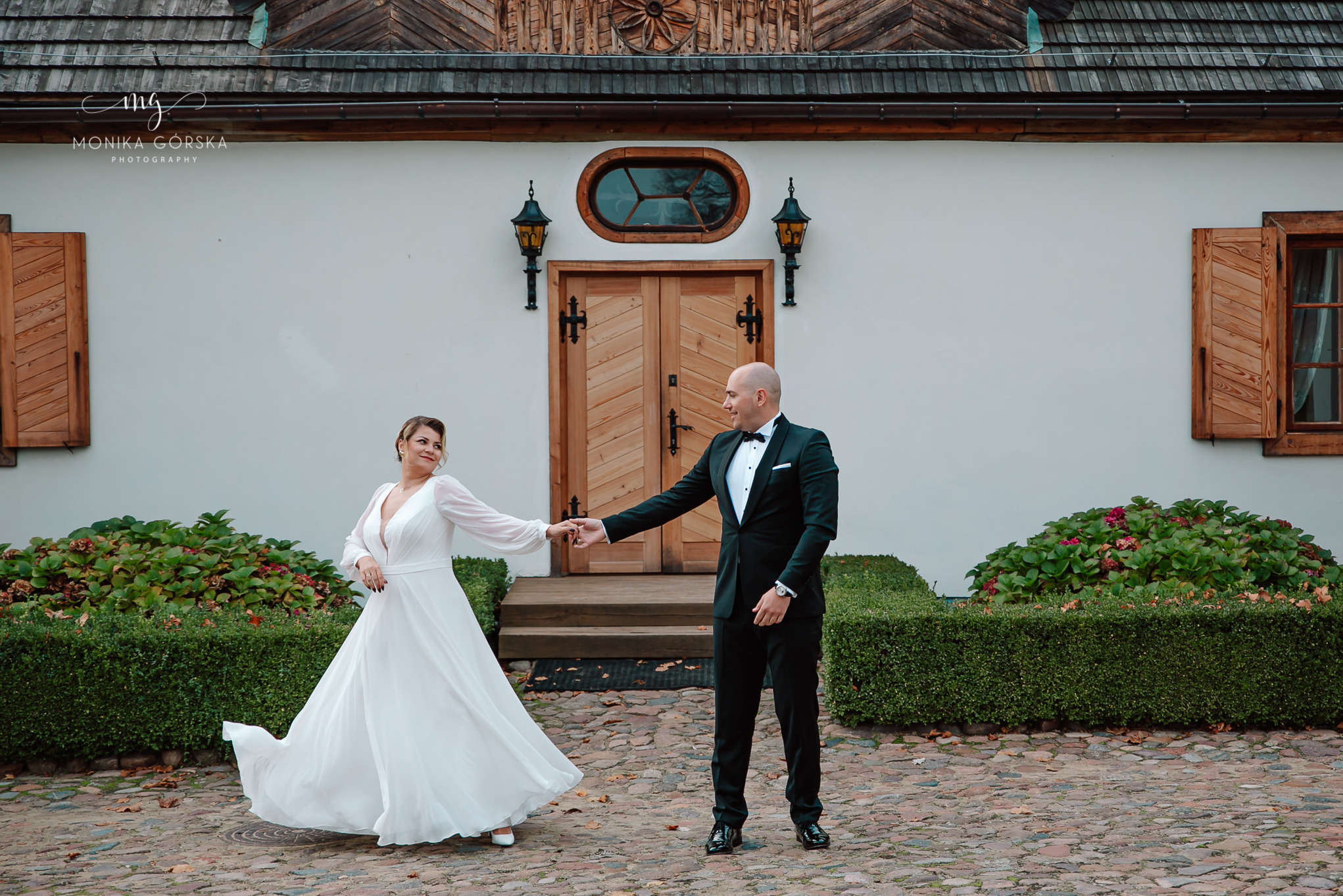 Para młoda trzymająca się za ręce na tle białego budynku z drewnianymi drzwiami i oknem. Ona w białej sukni, on w czarnym smokingu. Ujęcie z dołu, ciepłe światło.