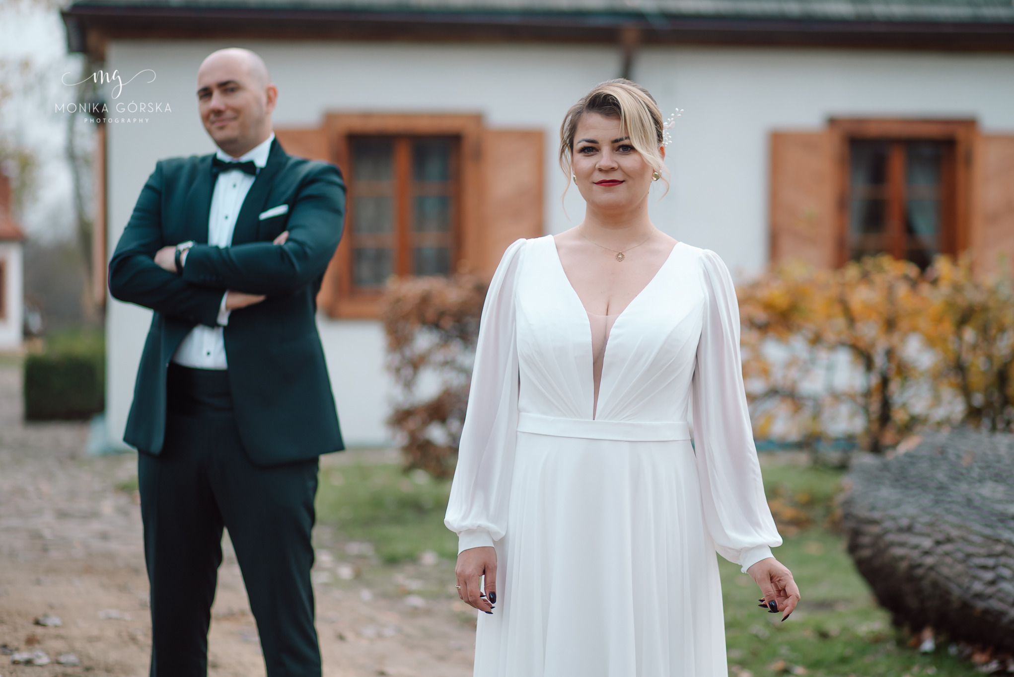 Elegancka para młoda: panna młoda w białej sukni z długimi rękawami, pan młody w czarnym smokingu, oboje na tle budynku z brązowymi okiennicami i jesiennych drzew.