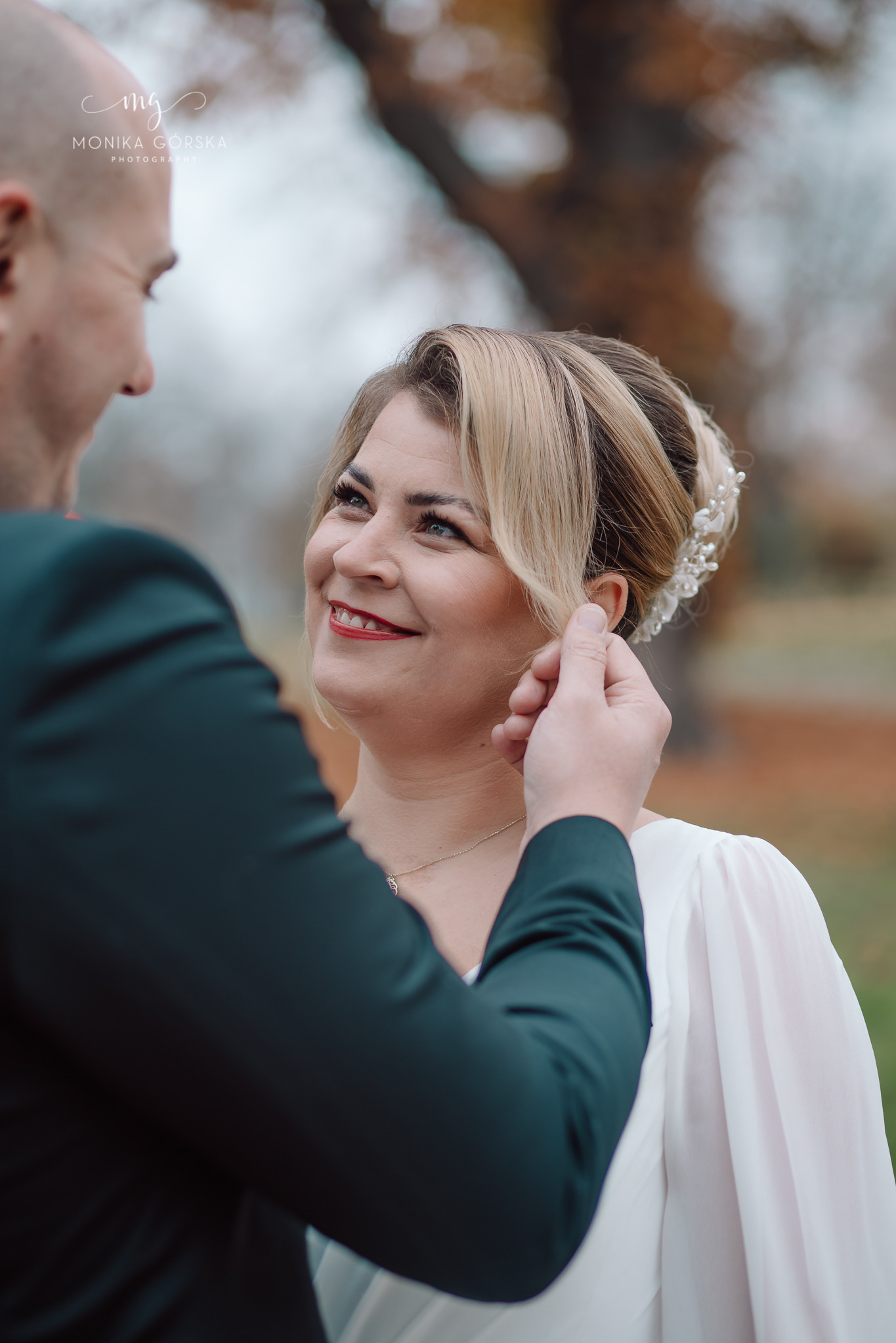 Uśmiechnięta panna młoda z blond włosami upiętymi perłami, w białej sukni, patrzy na pana młodego, który poprawia jej włosy. Jesienna sceneria w tle.