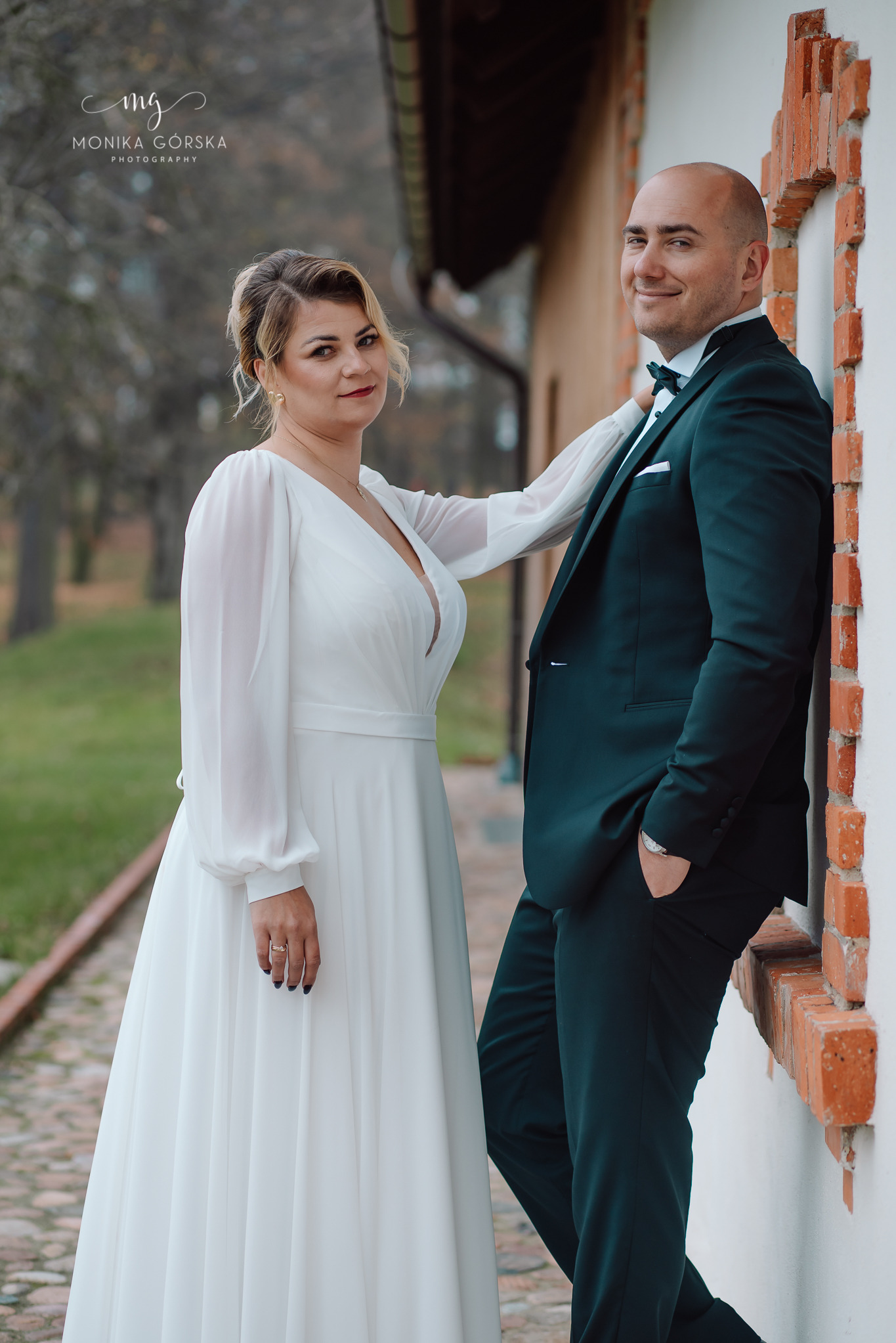 Elegancka para młoda: panna młoda w zwiewnej sukni i pan młody w butelkowej zieleni smokingu, pozują przy ceglanej wnęce okiennej w plenerze.