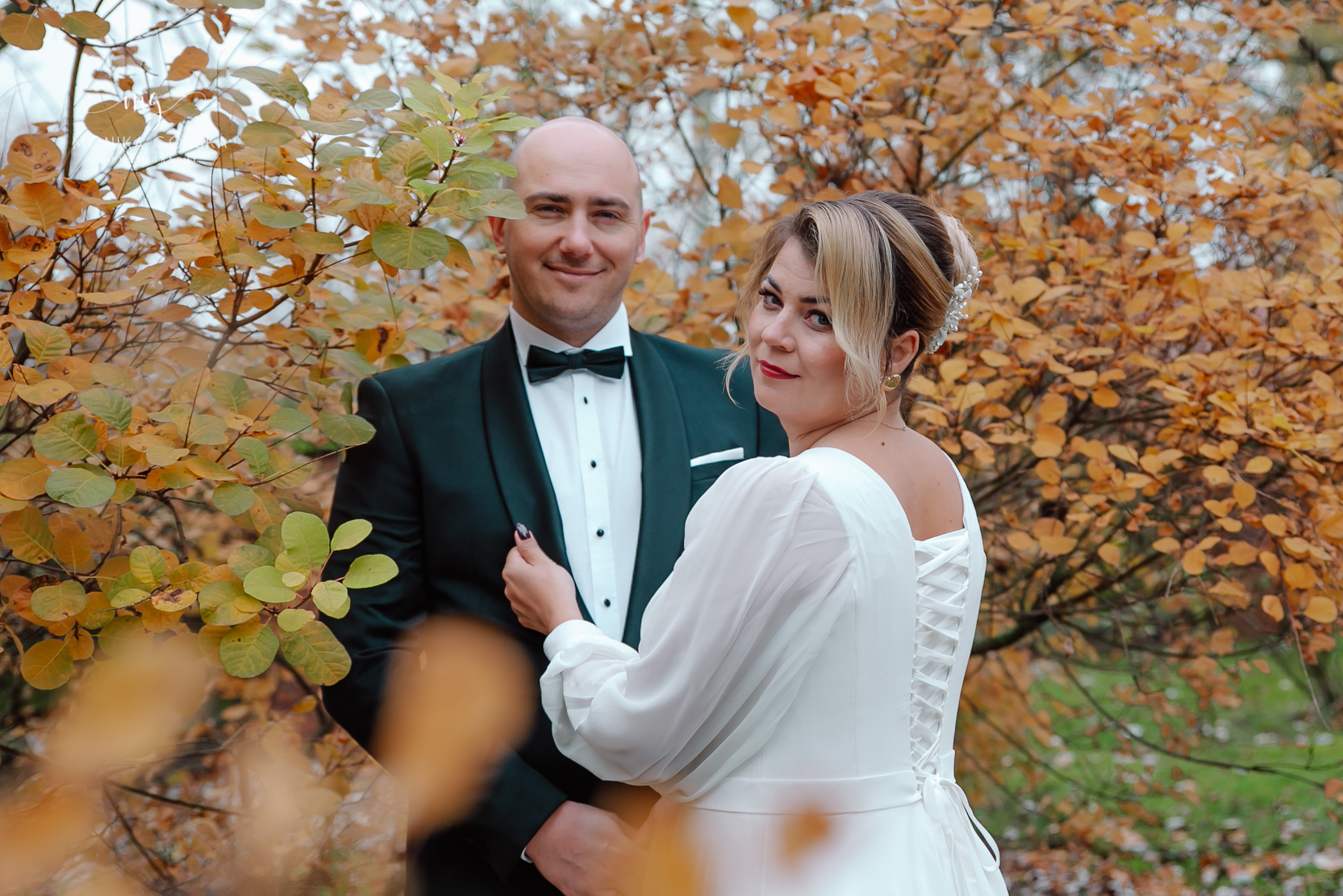 Uśmiechnięta para młoda w jesiennej scenerii: on w eleganckim smokingu, ona w białej sukni z koronkowym wiązaniem na plecach, pozują na tle drzew z żółtymi liśćmi.