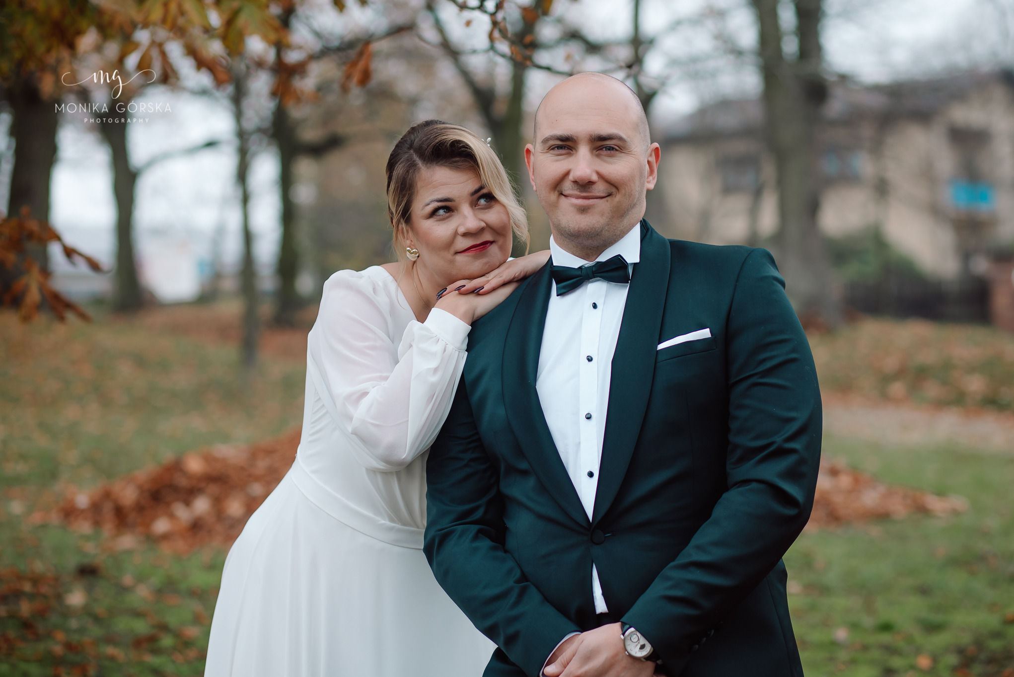 Uśmiechnięta para młoda w jesiennej scenerii: panna młoda opiera się o ramię pana młodego w ciemnozielonym smokingu. Romantyczna sesja w plenerze.