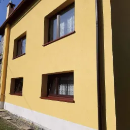 Żółta elewacja domu z brązowymi oknami i rynnami, widoczne fragmenty trawnika i kostki brukowej.