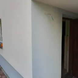 Jasnoszara elewacja budynku z widoczną fakturą tynku baranka, wystający przewód elektryczny przy wejściu, fragment okna z pomarańczowym elementem, oraz częściowo widoczny chodnik z kostki brukowej.