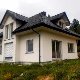 Wykończony dom jednorodzinny z ocieploną, białą elewacją, czarnym dachem z dachówki i częściowo opuszczonymi roletami zewnętrznymi na parterze, widok z poziomu gruntu.