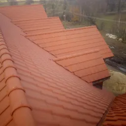 Nowy dach z czerwonej dachówki ceramicznej, widok z góry na fragment połaci z kominkiem wentylacyjnym, w tle jesienny krajobraz.