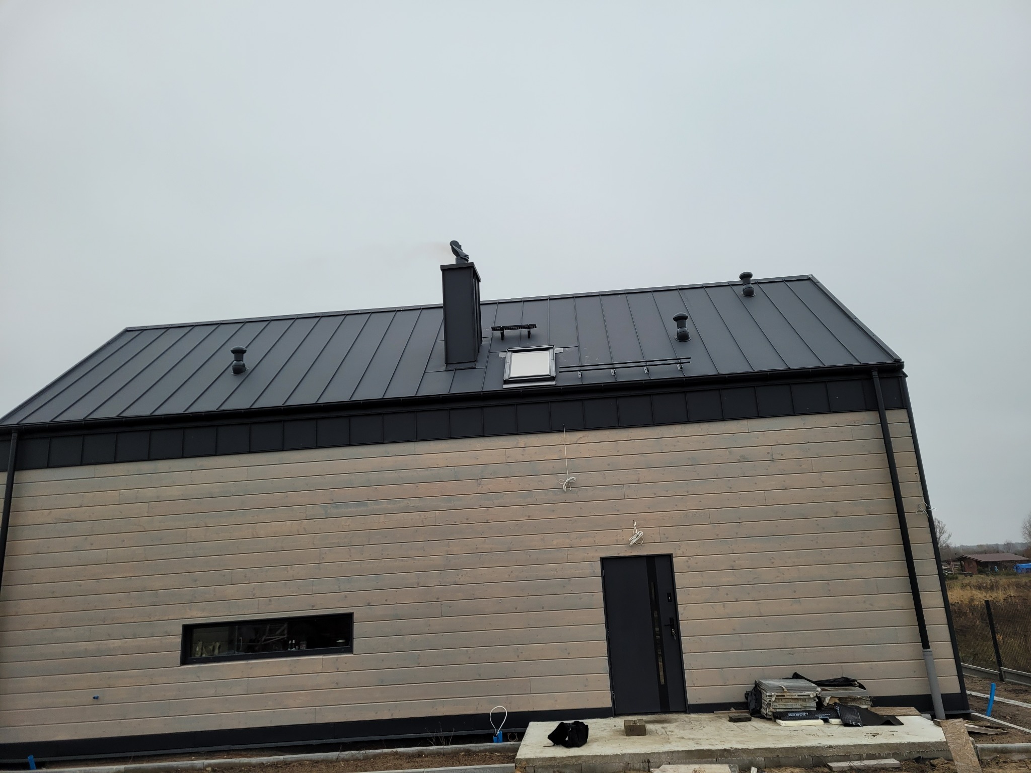 Nowoczesny dom z szarą, metalową dachówką, czarnym kominem z ozdobnym zakończeniem i niewielkim oknem dachowym. Ściany budynku wykończone drewnianą boazerią w odcieniach szarości i beżu.