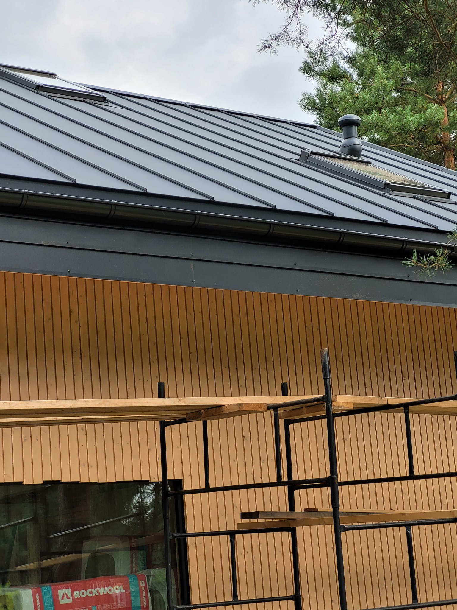 Czarny dach z blachy na domu z drewnianą elewacją, widoczne okna dachowe i komin wentylacyjny. Częściowo widoczne rusztowanie.