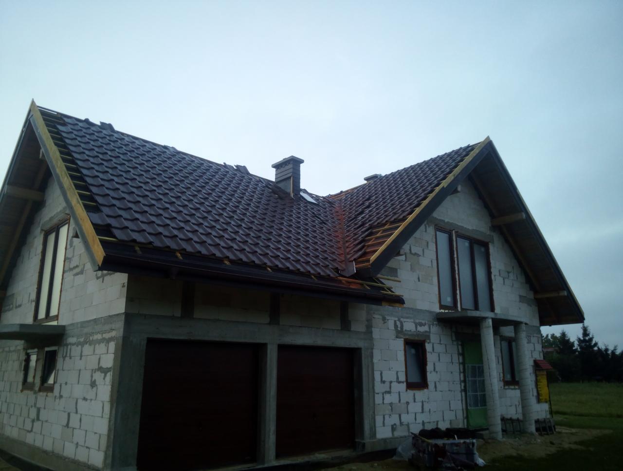 Dom w trakcie budowy z częściowo ułożonym, ciemnobrązowym dachem z dachówki modułowej. Widoczne elementy konstrukcji dachu oraz rynny. Ściany z białego bloczka, nieotynkowane. Okna w ciemnych ramach.