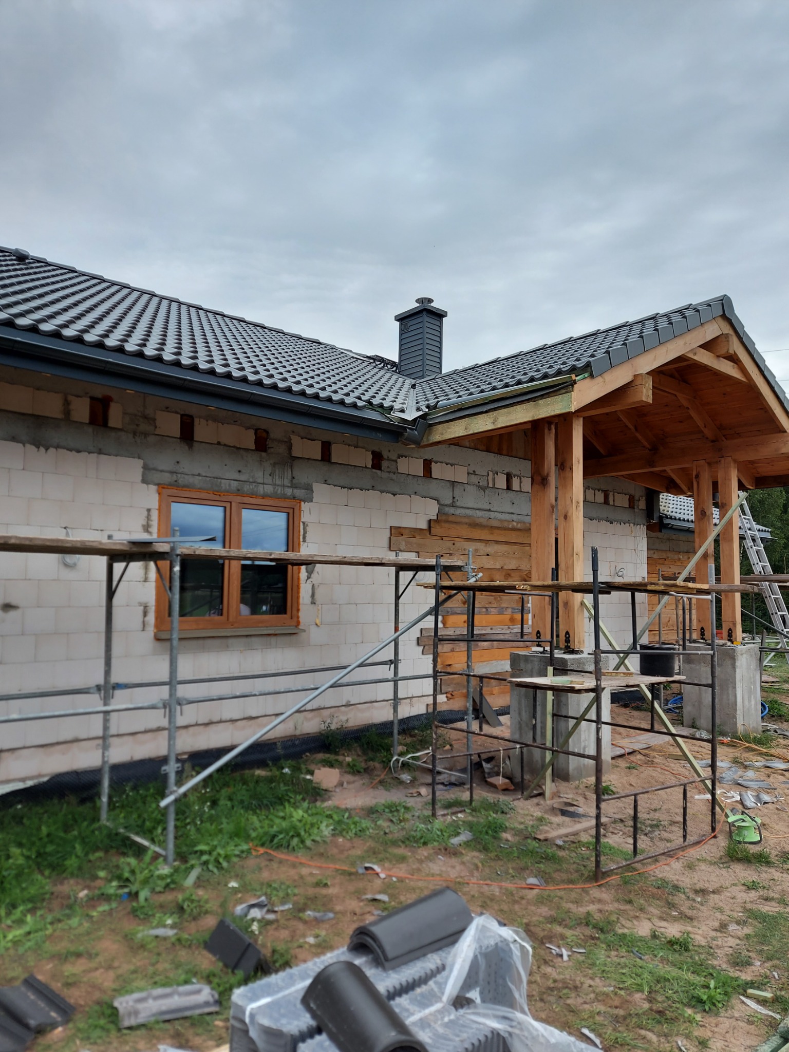 Dom w trakcie budowy, widoczne rusztowania, ściany z bloczków, drewniana konstrukcja zadaszenia wejścia i ułożony dach z czarnej dachówki karpiówki. Na pierwszym planie leżą zapakowane dachówki.