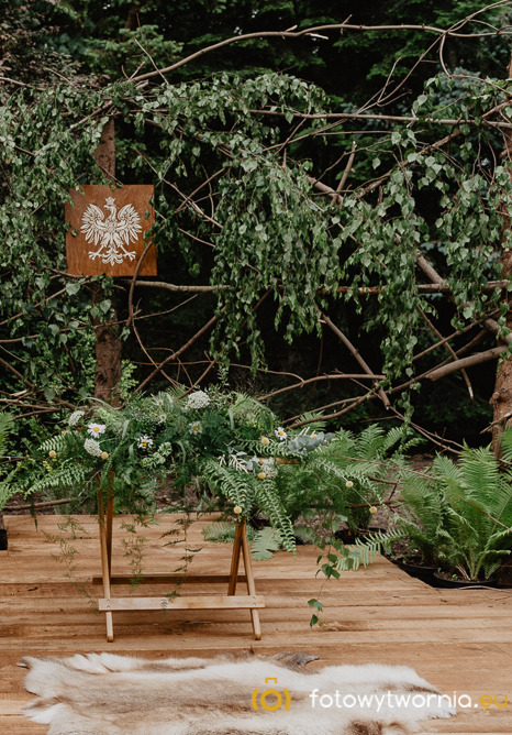 Dekoracja ślubna w stylu rustykalnym: drewniany stół udekorowany paprociami i białymi kwiatami, w tle drzewo z wygrawerowanym orłem w koronie na drewnianej tabliczce, na podłodze skóra.