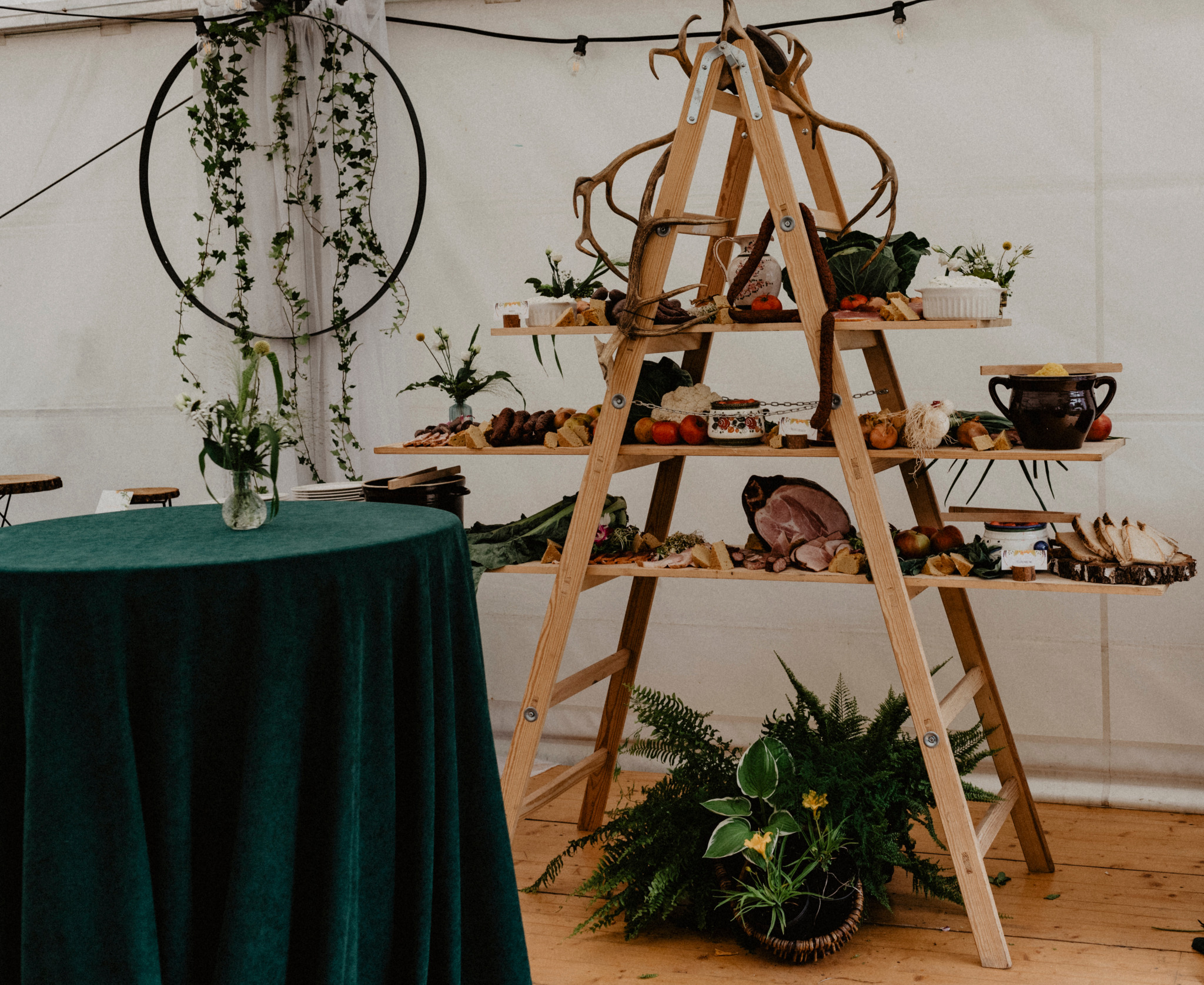 Rustykalny bufet na drewnianej drabinie z porożami, pełen wędlin, serów, owoców i warzyw, obok okrągły stół przykryty zielonym aksamitnym obrusem, dekoracje z bluszczu i kwiatów w tle.