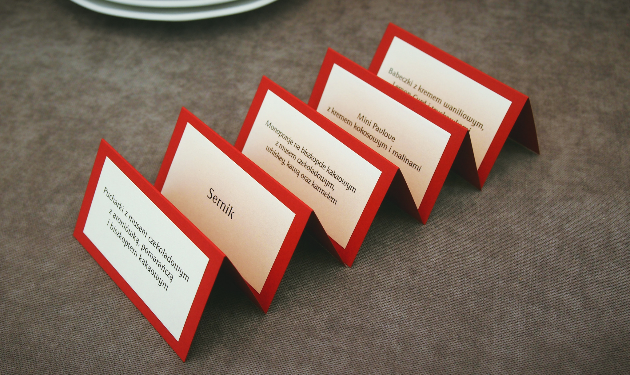 Rząd czerwono-białych winietek z nazwami deserów: Pucharki z musem czekoladowym, Sernik, Monoporcje, Mini Pavlove i Babeczki, ułożonych na szarym obrusie.
