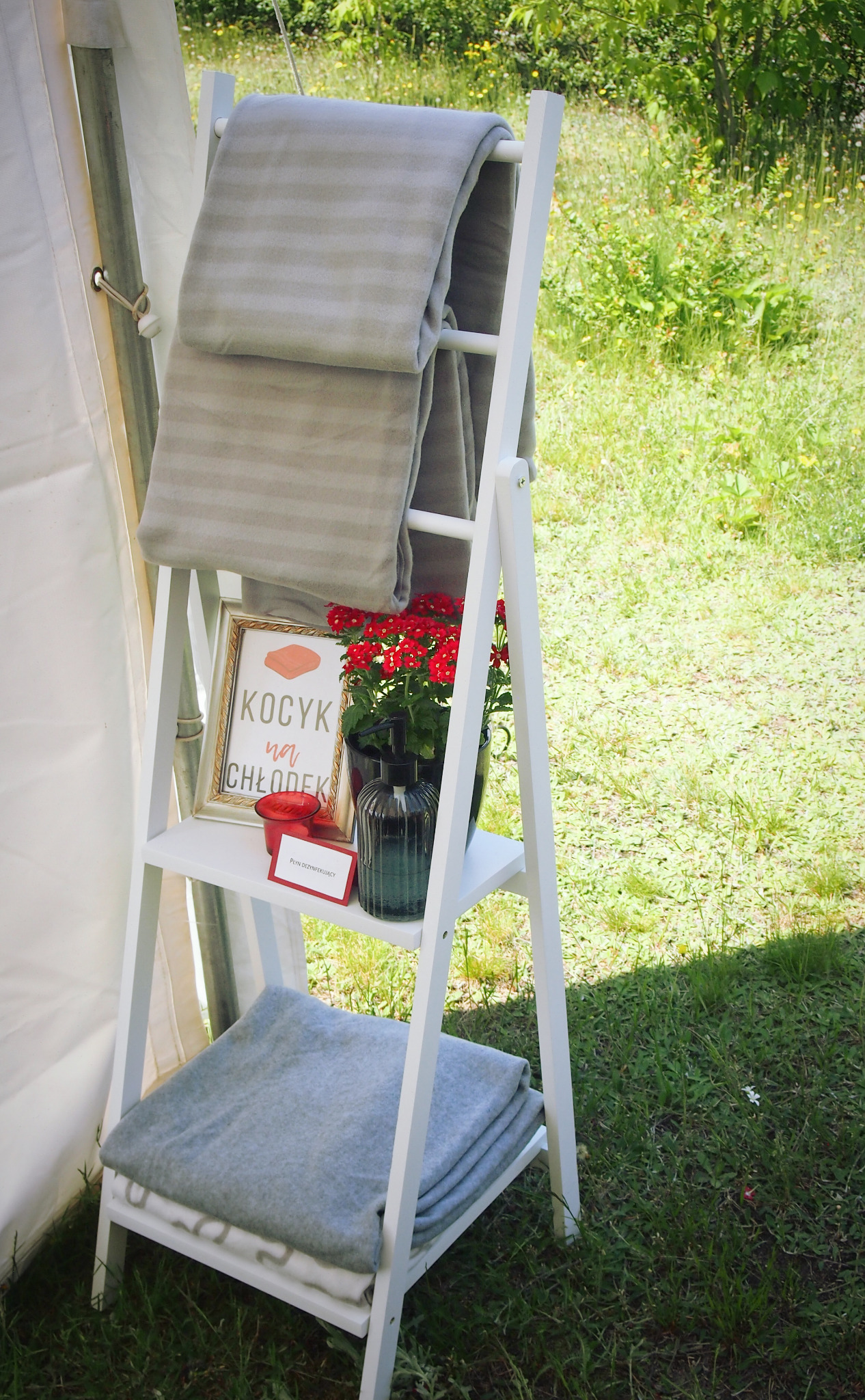 Biały, drewniany regał drabinkowy z szarymi kocami, czerwonymi kwiatami w czarnym wazonie, ramką z napisem 'Kocyk na chłodek' i butelką z dozownikiem płynu do dezynfekcji, ustawiony na trawie obok...