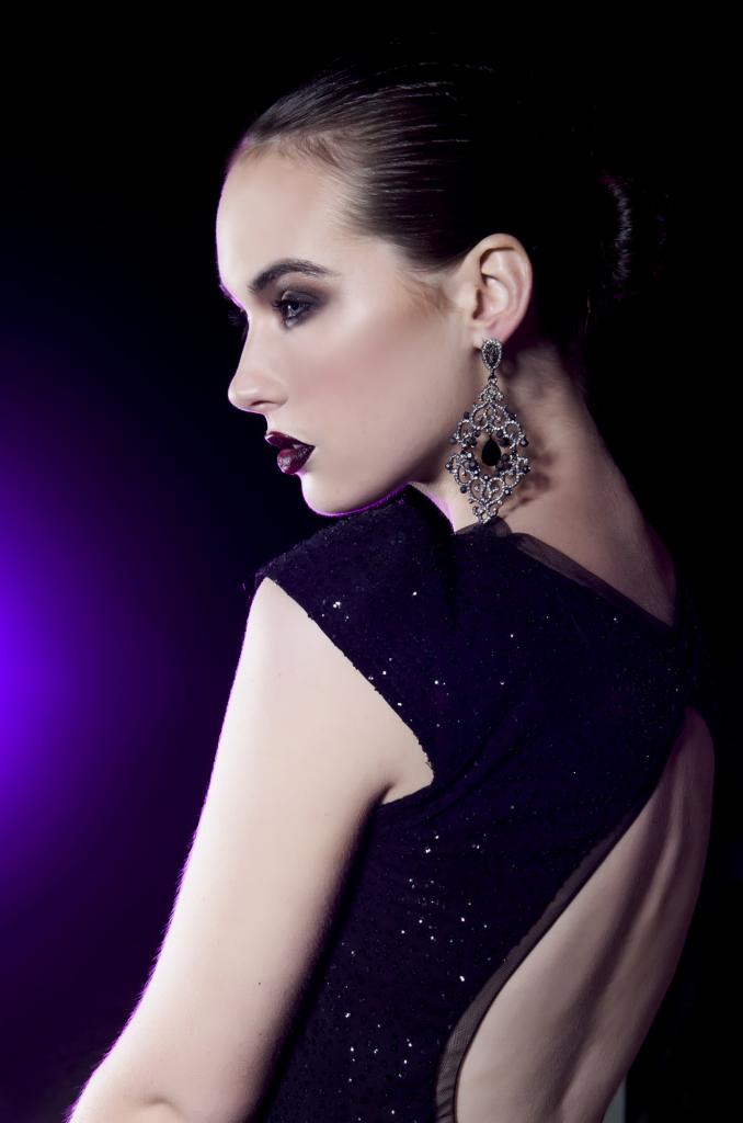 Profil kobiety z ciemną szminką, w czarnej sukni z cekinami i dużych, zdobnych kolczykach na czarnym tle z fioletowym akcentem.