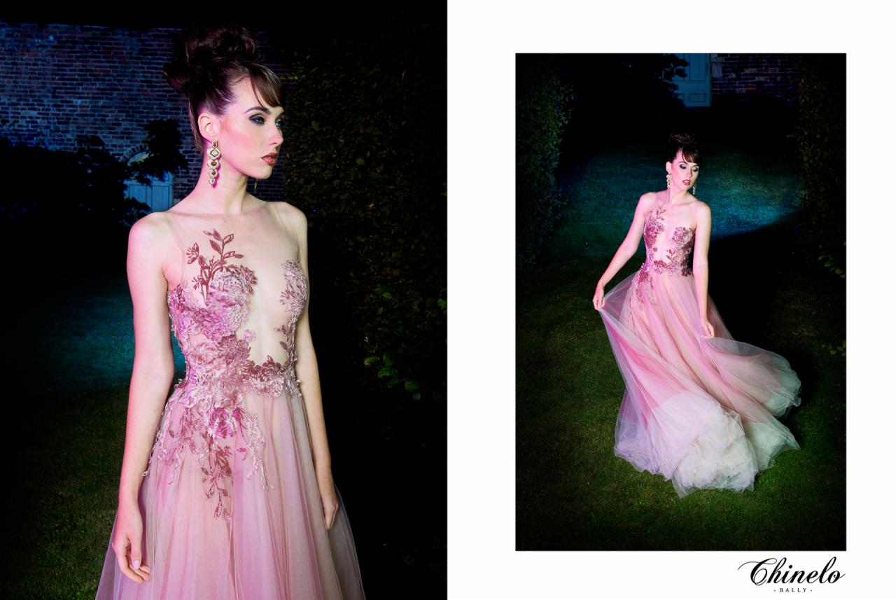 Kobieta w długiej, różowej sukni z kwiatowym haftem pozuje na tle zieleni i ceglanej ściany w nocnym oświetleniu, prezentując elegancką biżuterię.