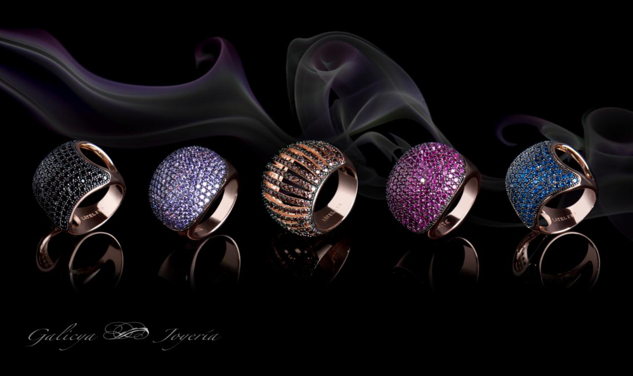 Pięć pierścionków z kolorowymi kamieniami na czarnym tle z efektem dymu w tle, każdy pierścionek ma inny kolor kamieni: czarny, fioletowy, brązowy, różowy i niebieski.