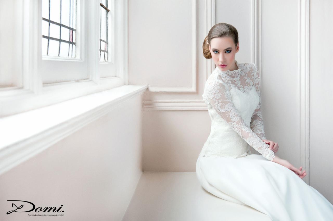 Panna młoda w koronkowej sukni z długim rękawem, siedząca na parapecie okna z widocznymi szprosami, w minimalistycznym wnętrzu oświetlonym naturalnym światłem.