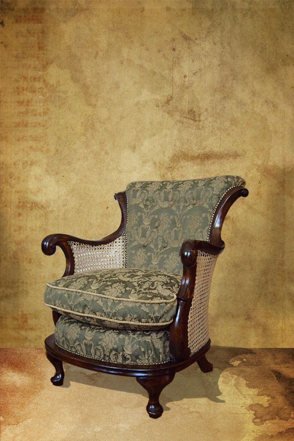 Antyczny fotel z ciemnego drewna, z zieloną tapicerką w kwiatowy wzór i rattanowymi bokami, na tle teksturowanej ściany w odcieniach beżu i brązu.