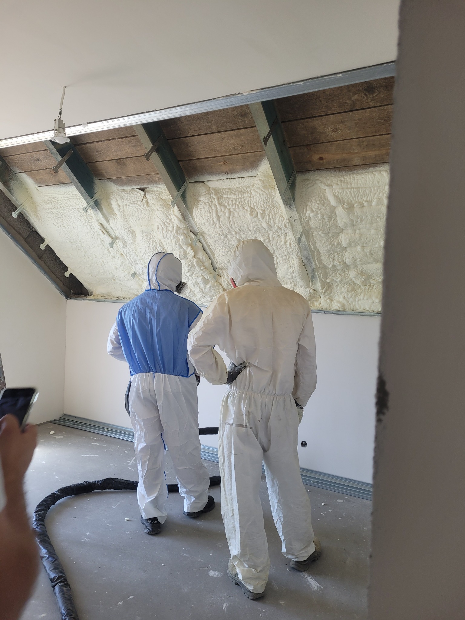Dwóch pracowników w kombinezonach ochronnych, w tym jeden z niebieską kamizelką, podczas aplikacji izolacji w poddaszu, widoczne drewniane krokwie i biała pianka izolacyjna.