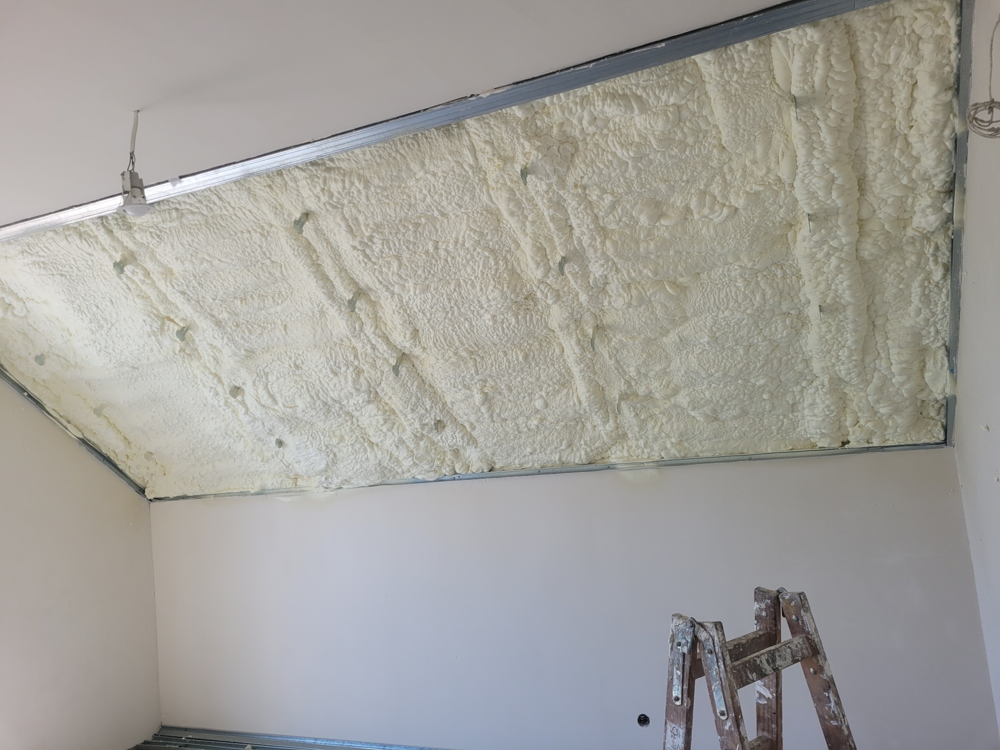 Pokrycie stropu jasną pianą izolacyjną, widoczne metalowe profile konstrukcyjne i fragment drabiny malarskiej w narożniku pomieszczenia.