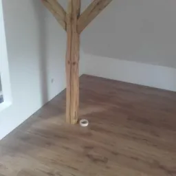 Poddasze z drewnianą belką konstrukcyjną, świeżo położone panele podłogowe w odcieniach brązu i białe ściany.