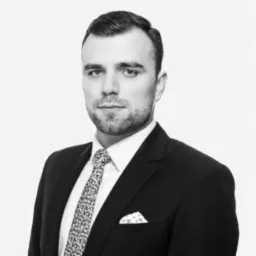 Portret biznesowy mężczyzny w czarnym garniturze i wzorzystym krawacie, na jasnym tle. Zdjęcie w odcieniach szarości, z wyraźnym światłem padającym z przodu.