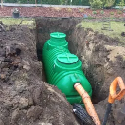 Zielony zbiornik z rurą w wykopie, widoczne czarne kable i łopata z pomarańczowym uchwytem. W tle trawnik i tuje.